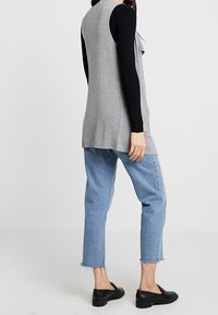 Szary bezrękawnik na czarną prążkowaną bluzkę z długim rękawem, w połączeniu z jasnoniebieskimi, skróconymi dżinsami i czarnymi butami wsuwanymi.