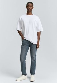 Weißes Baumwoll-T-Shirt mit kurzen Ärmeln, kombiniert mit schmal geschnittenen blauen Jeans und weißen Turnschuhen, stehend vor einem hellgrauen Hintergrund.