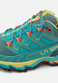 Zapatilla de correr turquesa con parte superior de malla, acentos amarillos y rojos, suela con agarre texturizado, sistema de cordones prominente y talón que ofrece soporte.