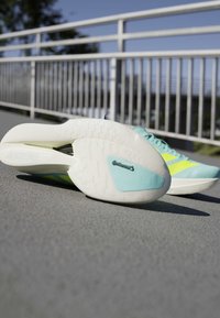 Lyseblå og gule løbesko med hvide såler, en sko viser Continental-logoet på sålen, placeret på en baneoverflade.