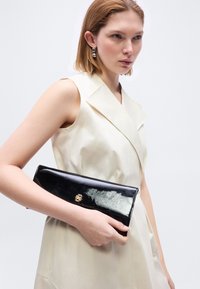 Γυναίκα με αμάνικο μπεζ φόρεμα που κρατά λαμπερό μαύρο τσάντα χειρός με χρυσό λογότυπο, φοράει ασημένια κρίκους σκουλαρίκια, σε απλό φωτεινό φόντο.