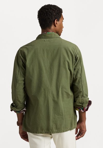 Camisa de manga longa verde oliva, com uma textura suave, colarinho aberto e mangas arregaçadas que revelam um forro estampado. A parte de trás apresenta um detalhe bordado.