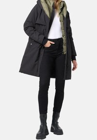 Svart parkajacka med huva, med en olivgrön foder, två bröstfickor och dragkedja. Bärs över svarta byxor och kraftiga stövlar.