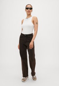 Bodysuit sin mangas blanco, jeans rectos marrón oscuro, gafas de sol negras, zapatillas blancas y marrones, y un collar de cuentas con un colgante.