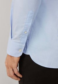 Camisa de botones azul claro con un tejido texturizado, puño puntiagudo y dos botones de perla en la manga. El dobladillo tiene una forma curva en la parte inferior.