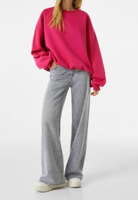 Sudadera fucsia de gran tamaño con hombros caídos, combinada con jeans de pierna ancha en gris claro y zapatillas de plataforma en crema. Texturas de tela suaves y casuales.