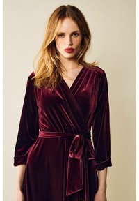 Burgundy sametti kietaisumekko, jossa on pitkät hihat, syvä V-pääntie ja sidottu vyö, korostaen sileää ja ylellistä tekstiiliä.