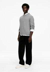 Grauer Strickpullover mit hohem Kragen, kombiniert mit schwarzen Cordhosen und weißen Sneakers. Das Modell steht vor einem einfarbigen Hintergrund.