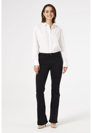 Femme debout, portant une chemise blanche boutonnée, un jean noir bootcut, des talons beiges et des boucles d'oreilles créoles, souriant à la caméra.