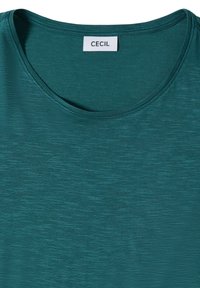 T-shirt col rond vert sarcelle avec un tissu doux et légèrement texturé, et une étiquette blanche cousue à l'intérieur de l'encolure.