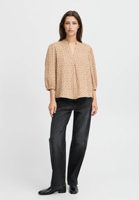 Beige Bluse mit V-Ausschnitt, verziert mit einem strukturierten Muster aus kleinen schwarzen Punkten, kombiniert mit dunklen, geraden Jeans und glänzenden schwarzen Schuhen.