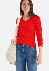 Top rosso a maniche lunghe con scollatura a V, realizzato in materiale morbido, abbinato a jeans blu. Il modello tiene una borsa di colore crema. Design semplice, senza pattern.