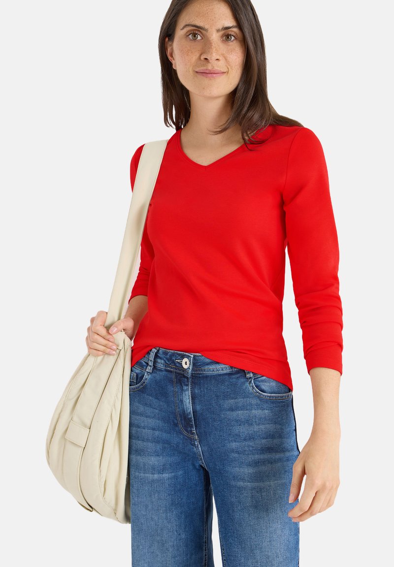 Top rosso a maniche lunghe con scollatura a V, realizzato in materiale morbido, abbinato a jeans blu. Il modello tiene una borsa di colore crema. Design semplice, senza pattern.