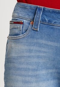 Närbild av en blå denimväv med synlig sömnad, en röd och vit etikett som sticker fram från framfickan, och en röd skjorta ovanför.