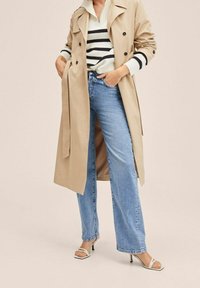 Woman wearing beige trench coat, white sweater with black stripes, light blue jeans, and white heeled sandals, hands in pockets.