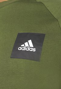 Materiał w kolorze oliwkowym o gładkiej fakturze z czarną prostokątną łatką z białym logo Adidas na środku.