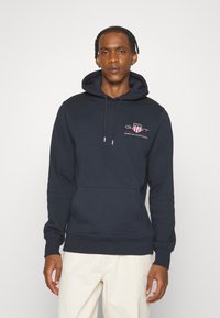 Marinblå hoodie med dragsko i huvan, framficka och en liten logotyp i rött och vitt. Mjuk bomullsblandning med ribbade ärmslut.