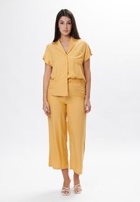Gele satijnen shirt met korte mouwen en een voorzak, gecombineerd met bijpassende broek met brede pijpen. De schoenen hebben een witte kruisriemontwerp.