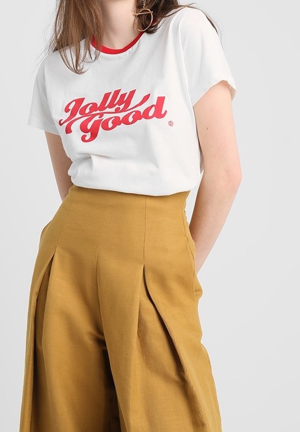 Personne portant un t-shirt blanc avec un texte rouge "Jolly Good" et un pantalon large taille haute couleur moutarde sur un fond uni.