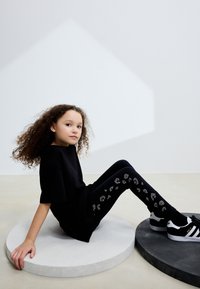 Črna majica s kratkimi rokavi skupaj s črnimi tajicami z srebrnimi okraski. Model sedi na svetlih in temnih okroglih platformah.