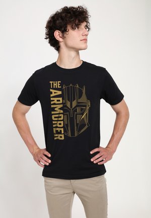 Junger Mann mit lockigem Haar, der ein schwarzes T-Shirt mit einem goldenen, stilisierten gepanzerten Helm und den Worten "THE ARMORER" trägt.