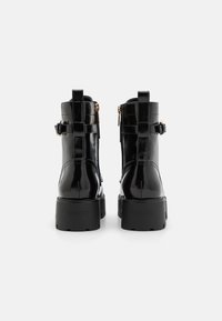 Bottines en cuir verni noir avec talons cubiques, fermetures éclair latérales et sangles décoratives. Elles présentent une texture lisse et un logo de marque embossé.