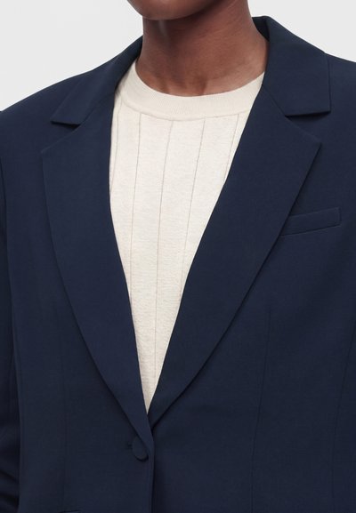 Personne portant un blazer bleu marine sur un pull crème côtelé à col rond, raccourci au niveau du cou et des épaules.