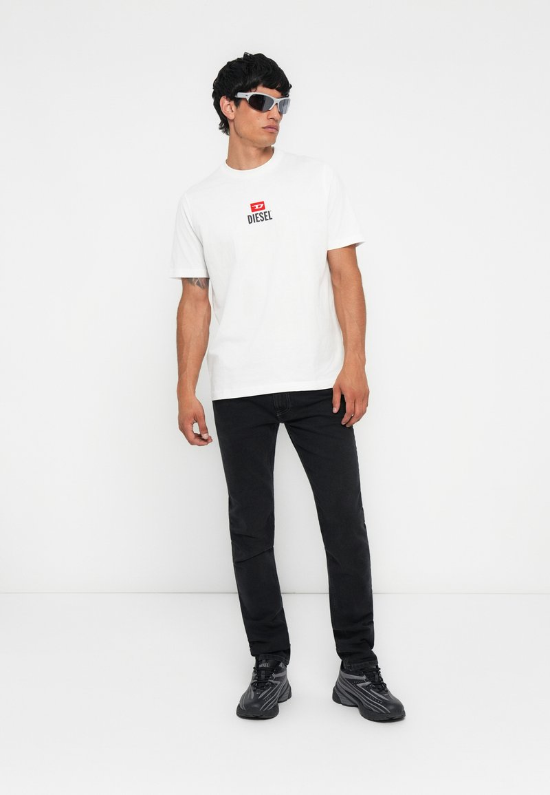 Mann trägt ein weißes Diesel-T-Shirt, schwarze Jeans, schwarze Sneakers und silberne Sonnenbrille und steht vor einem weißen Hintergrund.