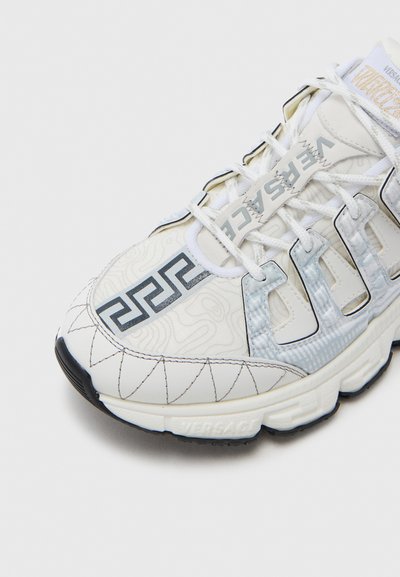 Baltas sportinis batelis su raštuota viršutine dalimi, mėlynomis detalėmis, siuvinėtu dizainu ir gumine padu. Turi raištelių užsegimą ir firminį ženklą.