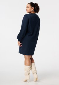 Robe tricotée bleu marine avec de longues manches bouffantes et des fentes latérales, associée à des bottes beiges décontractées jusqu'aux genoux, vue de dos.