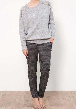 Femme portant un pull gris clair avec des découpes aux épaules, un pantalon slim à carreaux gris, et des talons ouverts transparents, debout sur un sol en bois.