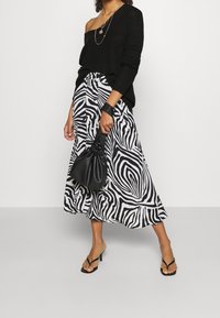Maglione nero a maglia con spalle scoperte abbinato a una lunga gonna a righe zebra in bianco e nero. Tiene una piccola borsa nera e indossa sandali neri con cinturini.