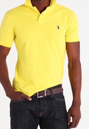 Homme portant un polo jaune vif à manches courtes et un jean foncé avec une ceinture marron, main dans la poche, sur un fond uni.