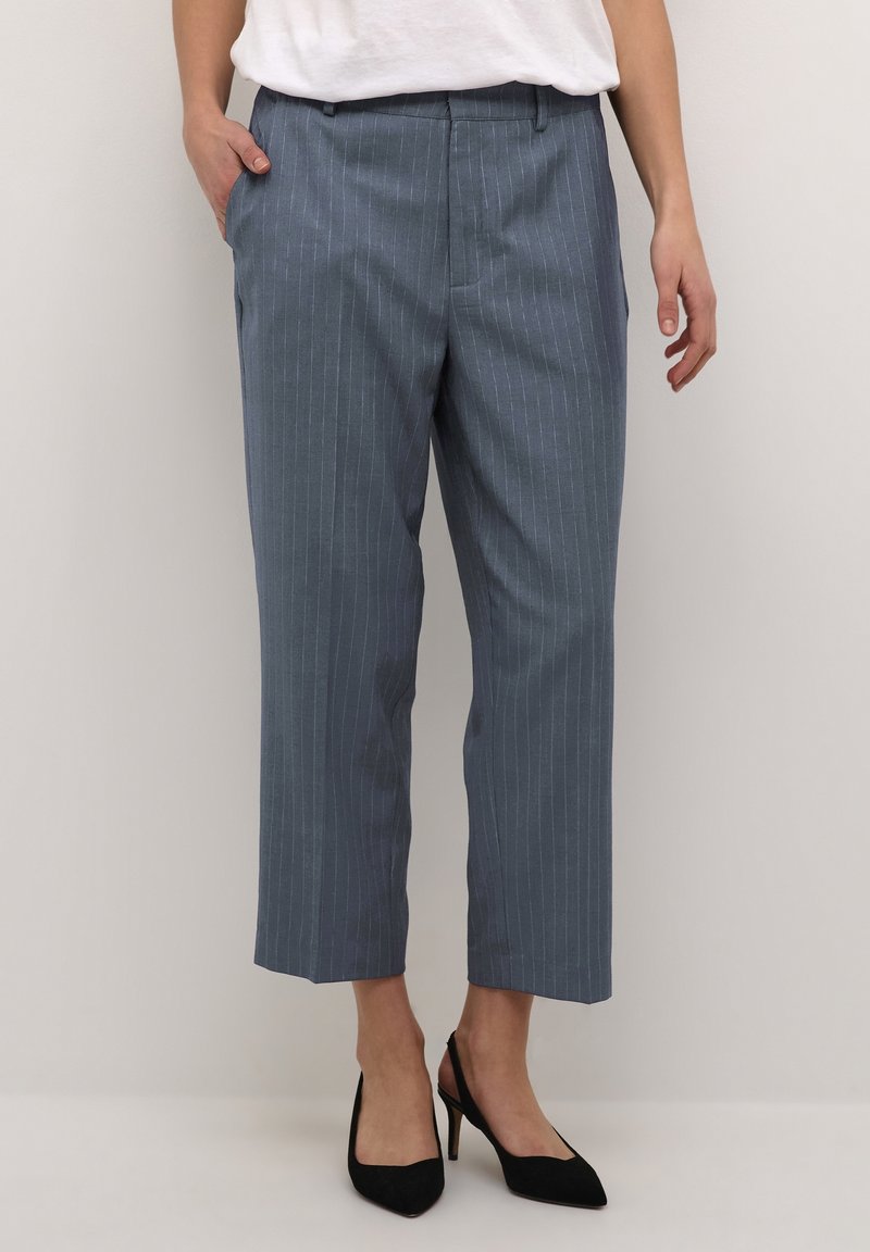 Kvinna som bär korta blå rutiga byxor med sidofickor och svarta högklackade slingback-skor, stående mot en enfärgad bakgrund.