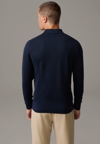 Navyblaues Langarm-Polo-Shirt mit Kragen, ausgestattet mit gerippten Bündchen und einer strukturierten Oberfläche. Getragen mit beigen Hosen.