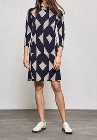 Robe à motif bleu marine et gris clair avec un design en losanges, manches trois-quarts et encolure ronde, associée à des baskets blanches.