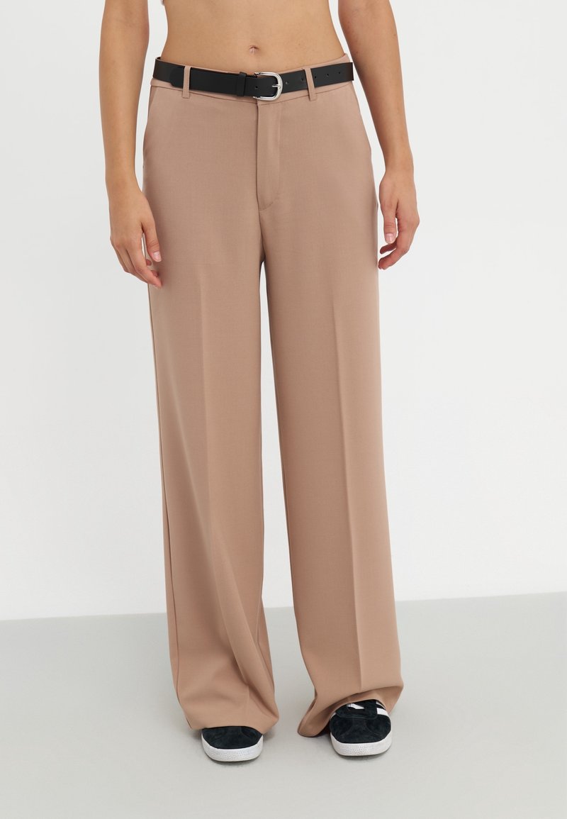 Pantalon beige à jambes larges avec ceinture noire, porté avec des baskets noires et blanches, présenté du torse aux pieds sur un fond uni.