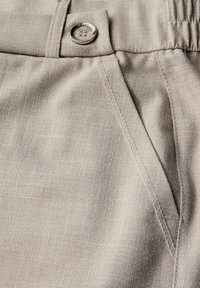 Pantaloni beige su misura realizzati in tessuto testurizzato. Presentano un bottone argento vicino alla vita e una tasca laterale. È visibile un sottile motivo diagonale.