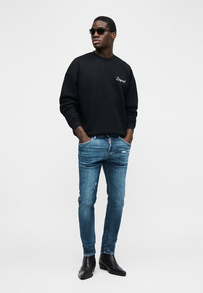 Άνδρας που φοράει μαύρο φούτερ, μπλε φθαρμένα τζιν, μαύρα δερμάτινα μποτάκια και σκούρα γυαλιά ηλίου, στέκεται με τα χέρια στις τσέπες.