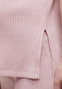 Haut en maille côtelée rose clair avec fente latérale, associé à un bas assorti en maille côtelée, laissant apparaître une partie d'une main sur le côté droit.