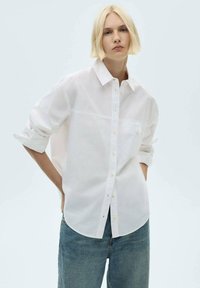 Camicia bianca con colletto a punta, maniche lunghe arrotolate, e una tasca sul petto; indossata con jeans blu a vita alta.