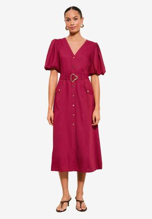 Žena v stredne dlhých fuchsiových šatách na gombíky s nafúknutými rukávmi, so zodpovedajúcim opaskom so srdcovou sponou a hnedými sandálmi na podpätku.
