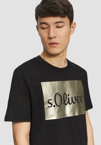 Zwart T-shirt met korte mouwen met een metallic gouden rechthoekige graphic met daarin "s.Oliver" in zwart cursief schrift. Klassieke ronde hals.