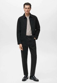 Črna denim jakna z dvema sprednjima žepoma, v kombinaciji s svetlo sivo majico in črnimi hlačami. Čevlji so sijajni črni usnjeni s zaobljenim nosom.