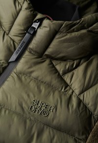 Giubbotto imbottito verde oliva con texture trapuntata, caratterizzato da un logo Superdry ricamato e una cerniera a contrasto.