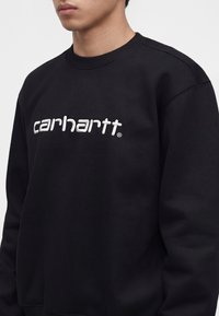 Junge Person trägt einen schwarzen Sweatshirt mit Rundhalsausschnitt, auf der Brust ist "carhartt" in Weiß gestickt.
