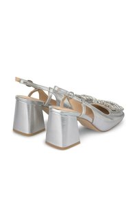 Scarpe metallizzate argento con tacco a blocco, caratterizzate da decorazioni sulla parte anteriore e cinturino regolabile alla caviglia. Texture liscia.