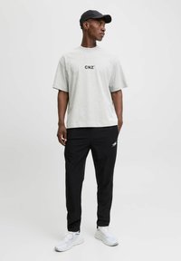 Mand iført en lysegrå oversized t-shirt med teksten "CNZ", sort kasket, sorte bukser med hvidt logo og hvide sneakers, står mod en hvid baggrund.