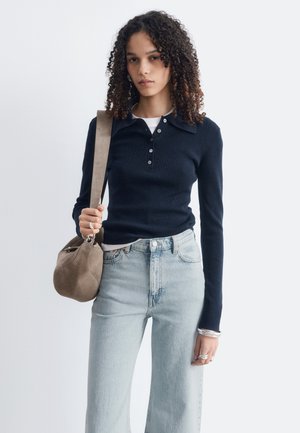 Jeune femme aux cheveux bouclés portant un polo noir à manches longues, un jean bleu clair, des bagues argentées, des bracelets et portant un sac à épaule en daim marron.
