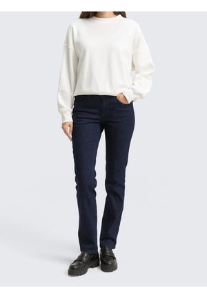 Femme portant un sweat-shirt blanc uni à manches longues, un jean skinny foncé et des mocassins noirs épais, debout avec une main dans la poche.